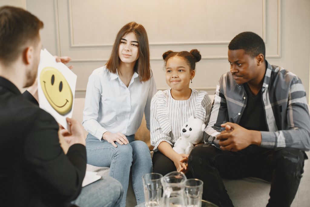 Image of mother, father, and child sitting on therapist couch with therapist sitting across from them and holding image of happy face.