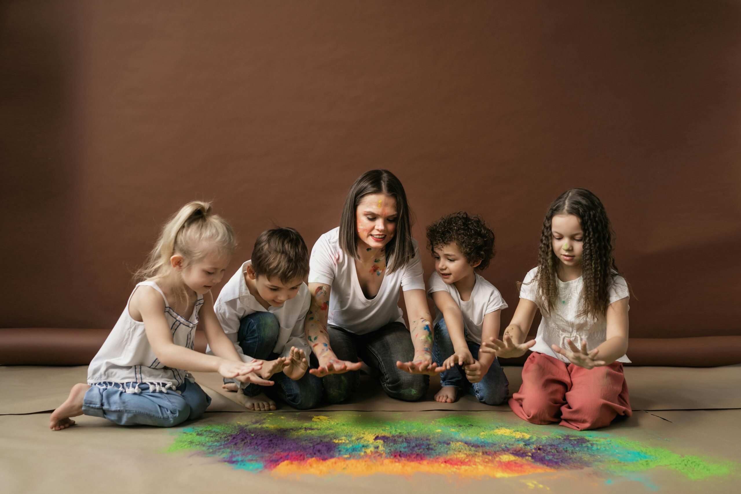 Image of adult on the ground with four young children as all are engaging in finger paint with each other.
