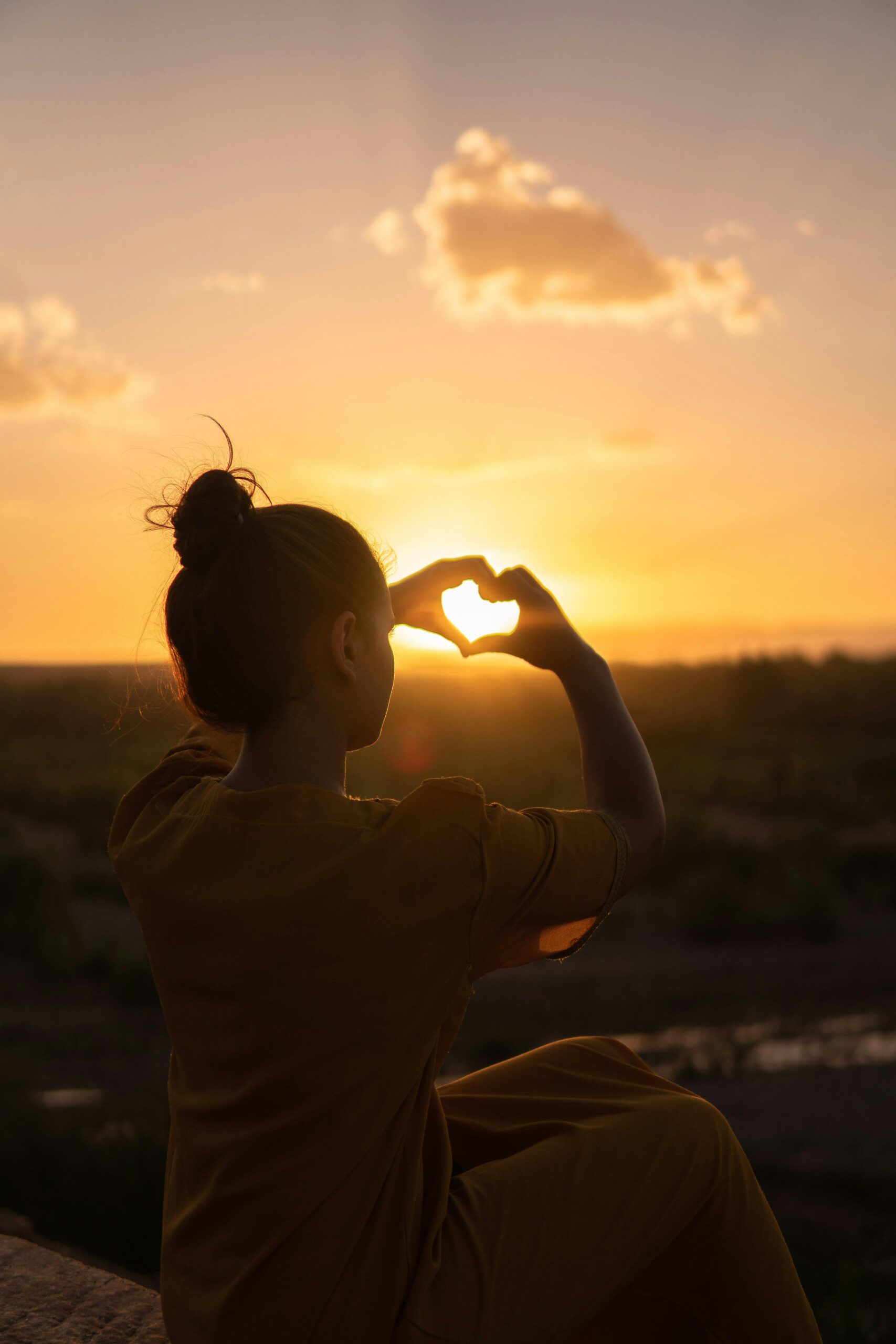 Image of woman forming shape of heart with fingers with a backdrop of the sunset.