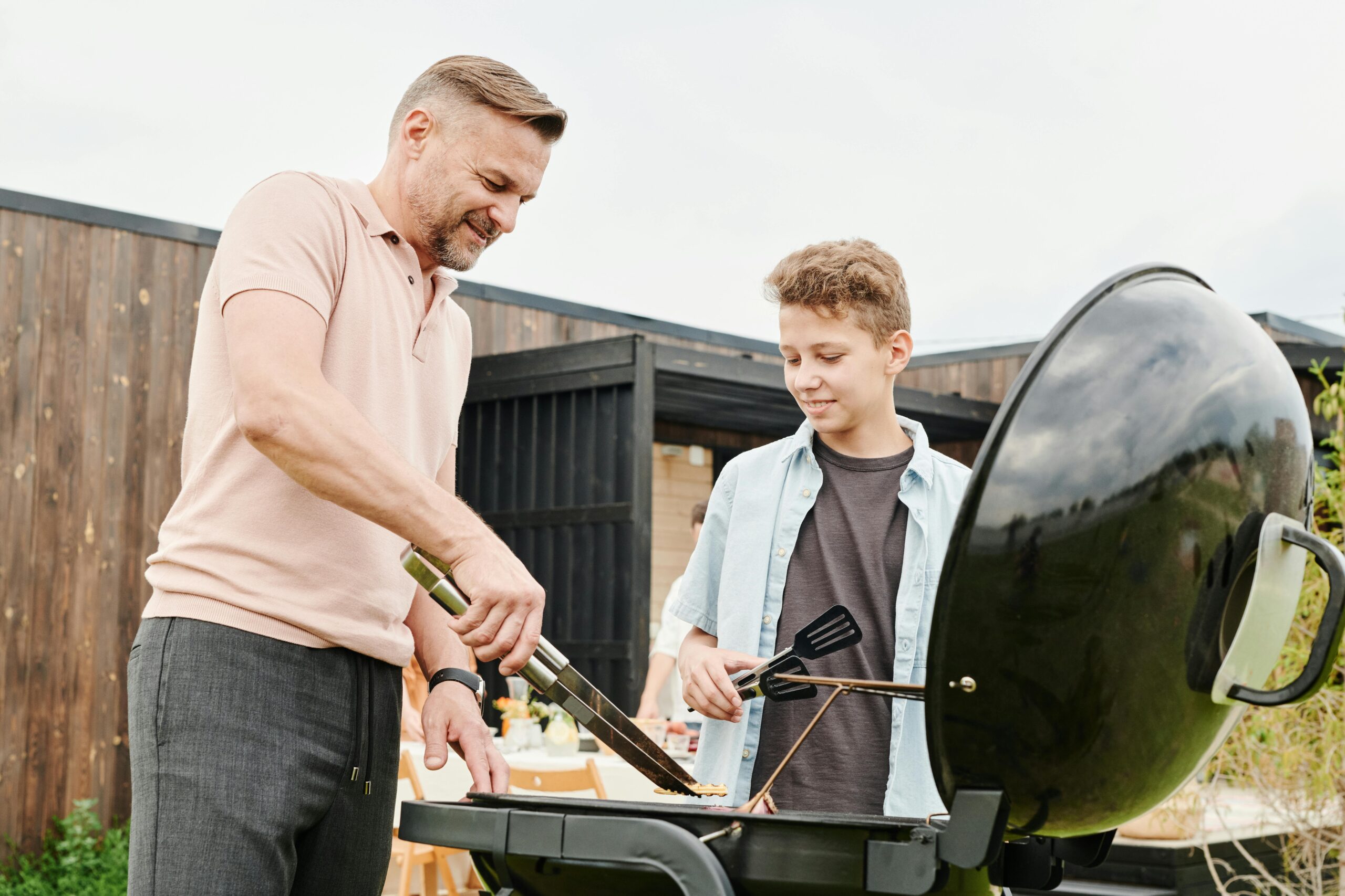 Image of father grilling food on BBQ outdoors with son helping.