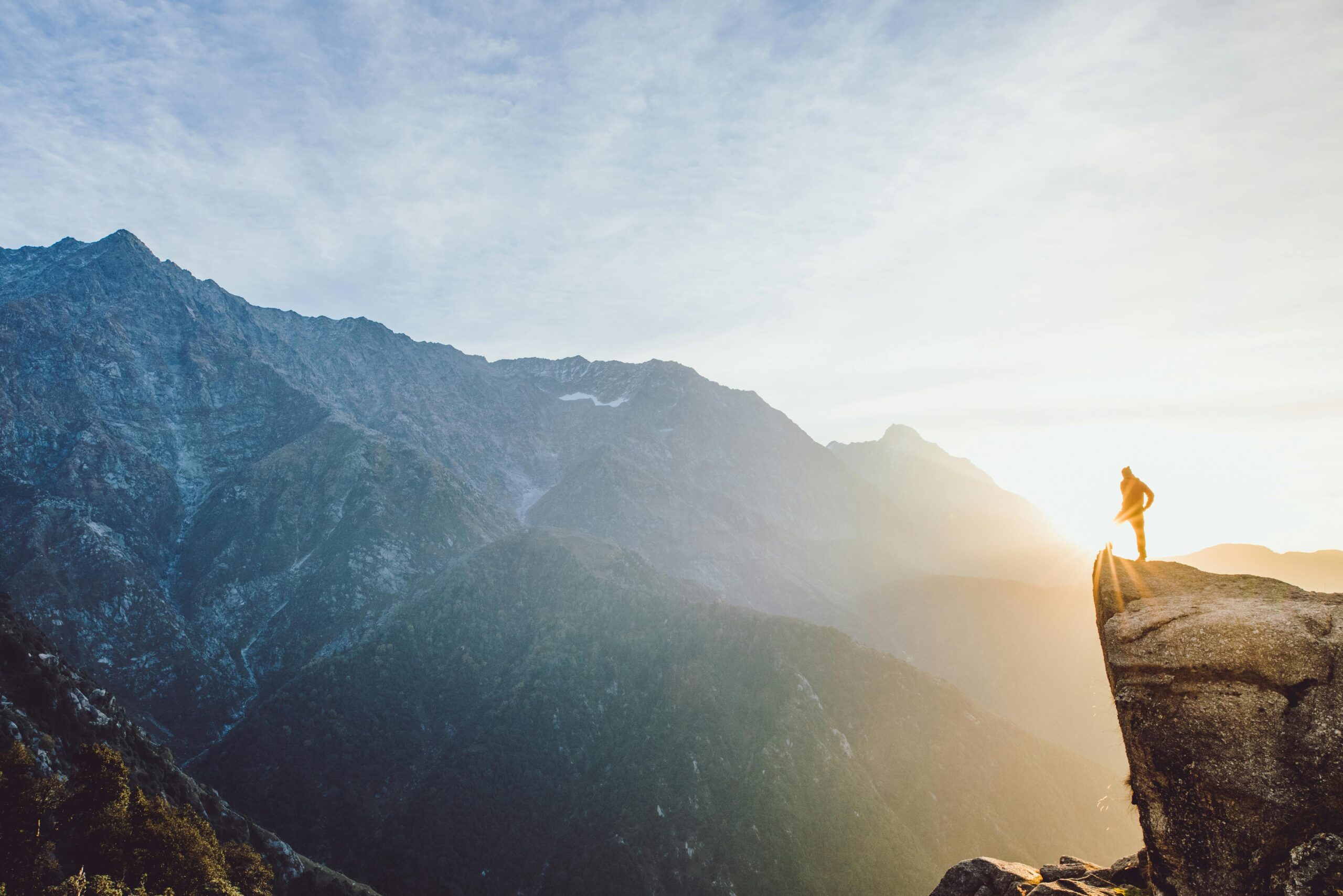 Image of person standing on mountain top looking out into the vast mountain range with the sun setting in background.