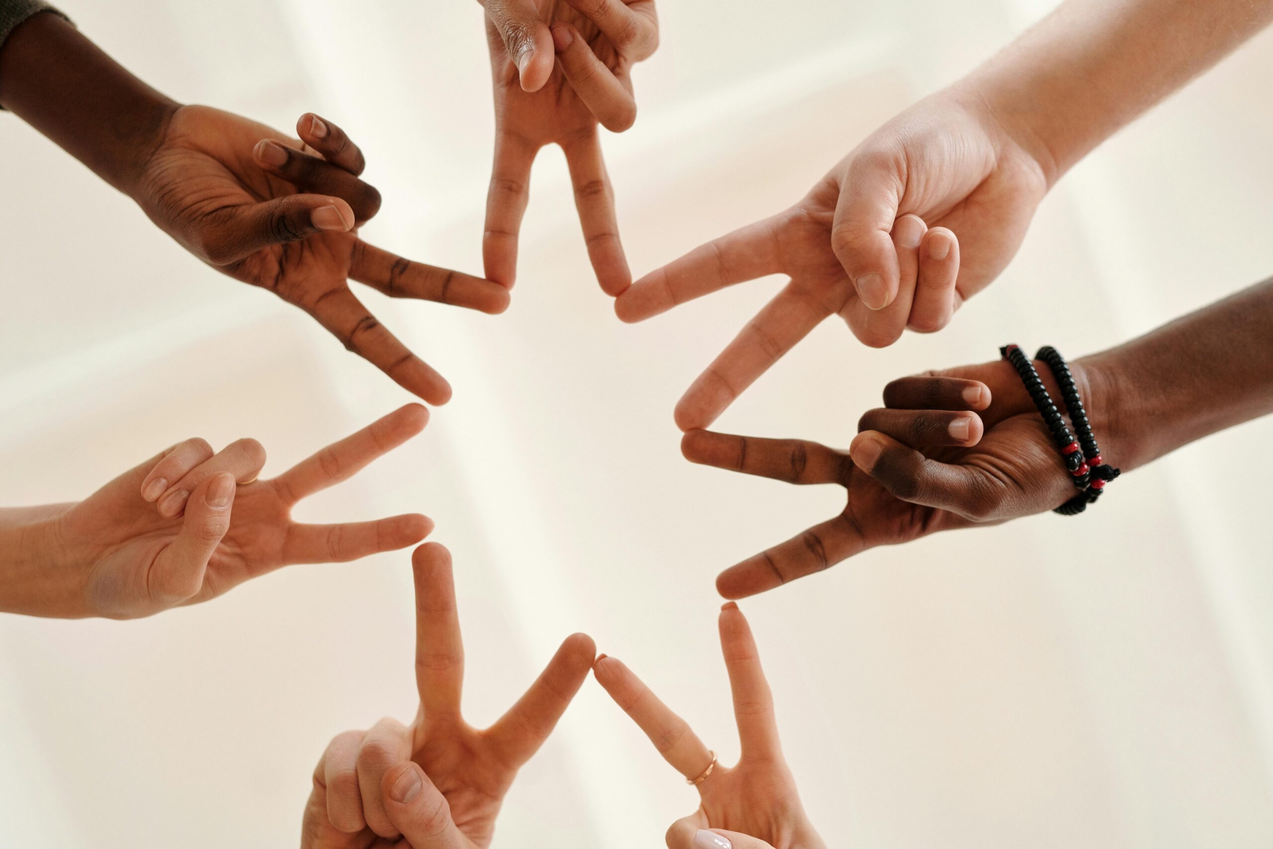 Image of seven hands each making the peace symbol with two fingers and all fingers connecting together with one another to form the shape of a star.