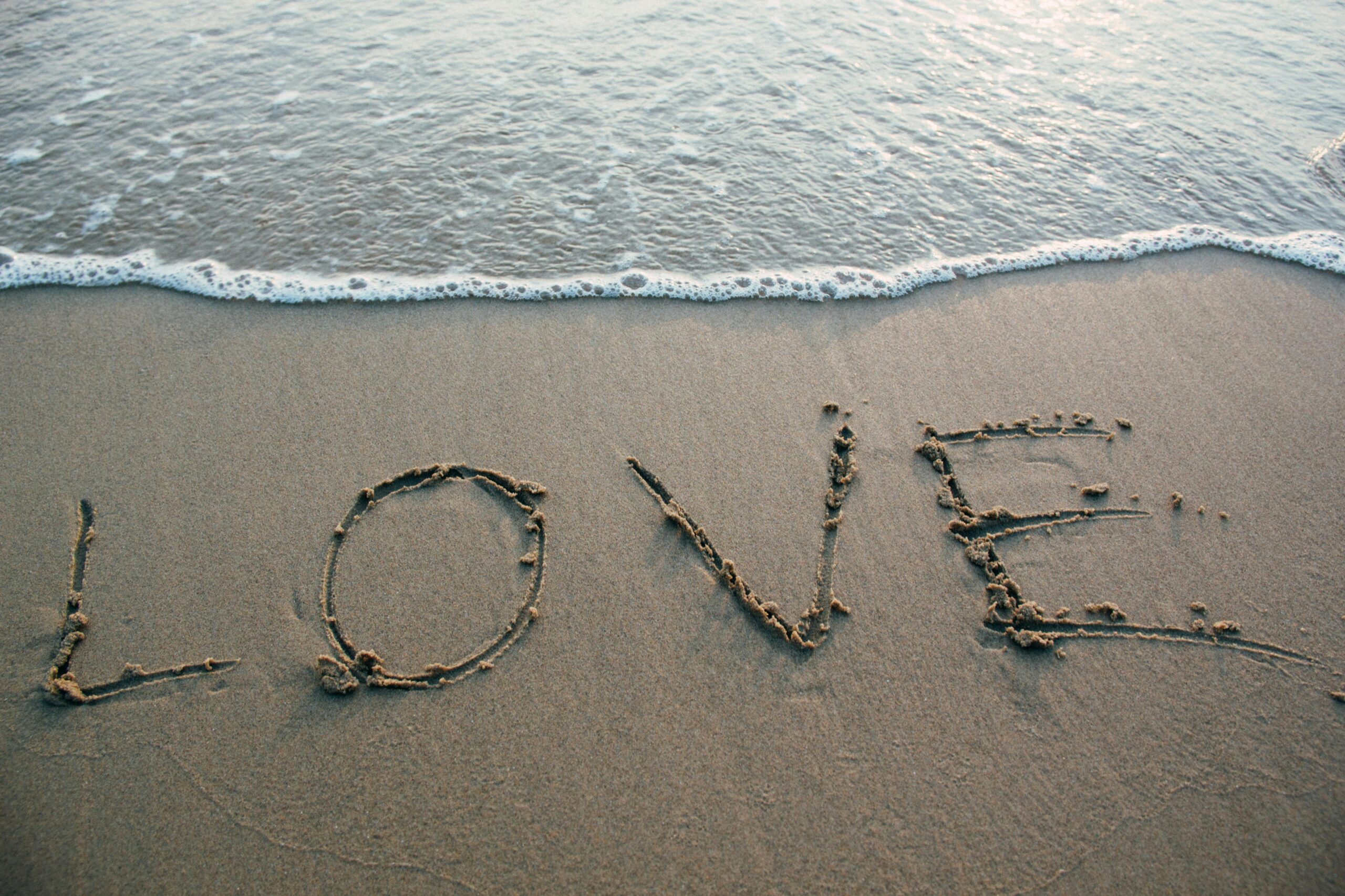 Image of beach with sand and ocean. The sand has the word "love" largely carved into it, as the backdrop to the words, "Couples Therapy."