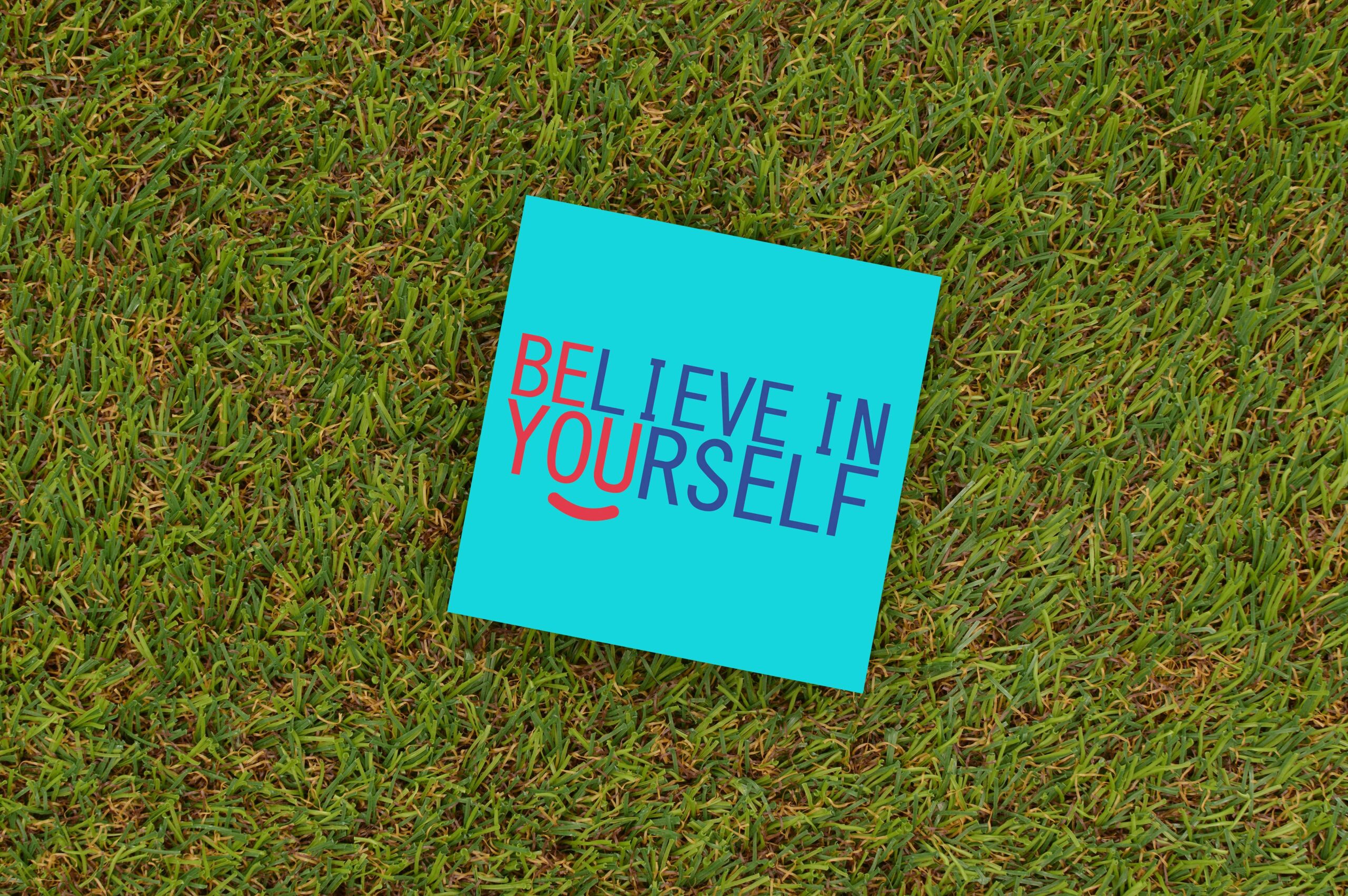 Image of a blue note laying on grass with the following phrase written on it, "Believe In Yourself," with the first letters in the color red that highlight the letters that spell out the following words: "Be You."
